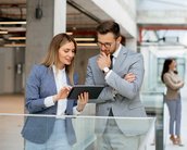 Ein Mann und eine Frau in formeller Kleidung betrachten gemeinsam ein Tablet in einem modernen Büro. Im Hintergrund sind weitere Personen zu sehen.