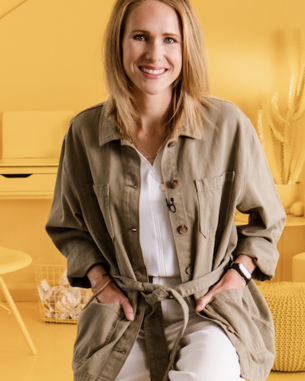 Eine Frau mit blonden Haaren trägt eine khakifarbene Jacke und sitzt in einem gelben Raum. Sie lächelt und hat eine weiße Bluse und helle Hosen an. Im Hintergrund sind minimalistische Möbel und Pflanzen sichtbar.