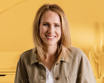 Eine Frau mit blonden Haaren trägt eine khakifarbene Jacke und sitzt in einem gelben Raum. Sie lächelt und hat eine weiße Bluse und helle Hosen an. Im Hintergrund sind minimalistische Möbel und Pflanzen sichtbar.