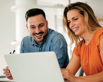 Zwei Personen sitzen an einem Tisch und schauen gemeinsam auf einen Laptop. Der Mann hat einen Bart und trägt ein blaues Hemd, die Frau hat lange Haare und trägt ein oranges Kleid.