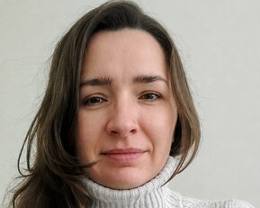 Eine Frau mit langen, braunen Haaren trägt einen hellen, gestrickten Rollkragenpullover. Sie schaut direkt in die Kamera.