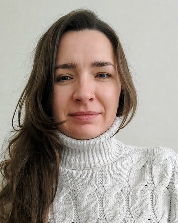Eine Frau mit langen, braunen Haaren trägt einen hellen, gestrickten Rollkragenpullover. Sie schaut direkt in die Kamera.