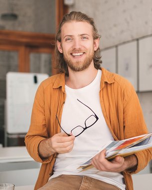 Ein junger Mann mit langen Haaren sitzt auf einem Tisch, hält eine Mappe mit Unterlagen und eine Brille in der Hand. Er trägt ein orangefarbenes Hemd und lächelt.