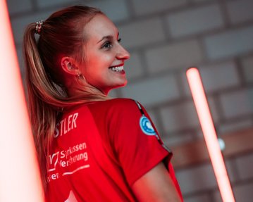 Eine lächelnde Frau trägt ein rotes Sporttrikot mit der Nummer 1. Ihr Haar ist lang und sie hat einen Pferdeschwanz. Im Hintergrund sind leuchtende Neonlichter zu sehen.