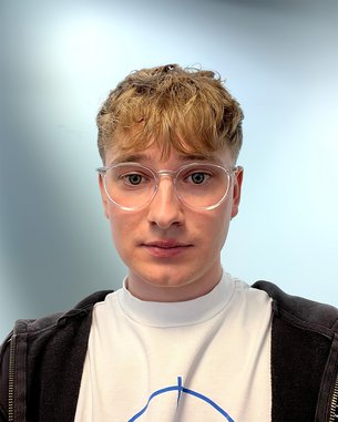 Junger Mann mit kurzen, lockigen Haaren und Brille, trägt ein weißes T-Shirt und einen schwarzen Hoodie, schaut direkt in die Kamera.