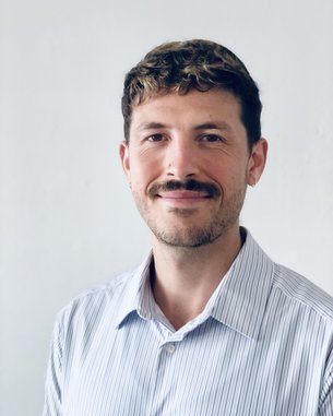 Ein Mann mit einem Schnurrbart trägt ein gestreiftes Hemd und lächelt in die Kamera. Der Hintergrund ist hell und neutral.