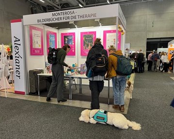 Messestand mit dem Namen "Monterosa" für Therapie-Bilderbücher. Zwei Personen stehen an einem Tisch, während ein Hund im Hintergrund liegt. Bunte Plakate hängen an der Wand.