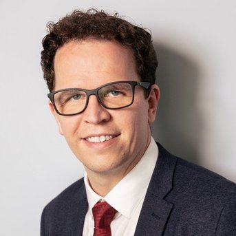 Ein Mann mit lockigem Haar und Brille trägt einen Anzug und ein Hemd mit Krawatte. Er lächelt leicht und schaut in die Kamera. Der Hintergrund ist hell und neutral.