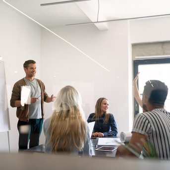Ein Mann präsentiert in einem Besprechungsraum, während mehrere Personen aufmerksam zuhören und Fragen stellen. An einer Wand steht ein Flipchart mit Diagrammen.