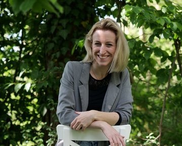 Eine Frau sitzt lässig auf einem weißen Hocker, trägt eine graue Jacke und Jeans, umgeben von grünen Pflanzen.