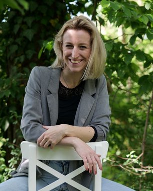 Eine Frau sitzt lässig auf einem weißen Hocker, trägt eine graue Jacke und Jeans, umgeben von grünen Pflanzen.