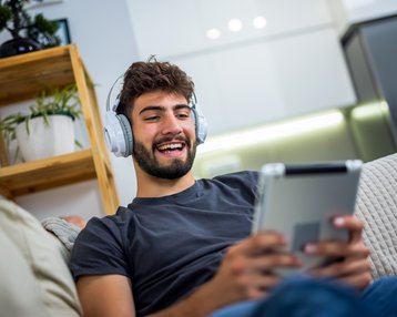 Junger Mann mit Kopfhörern, der auf einem Tablet sitzt und lächelt, während er entspannt auf einem Sofa ist.