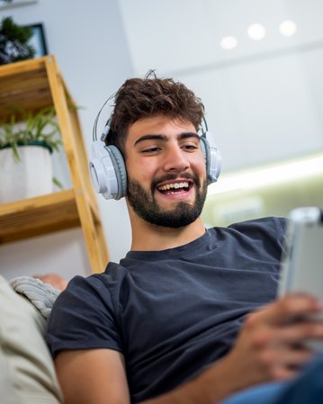 Junger Mann mit Kopfhörern, der auf einem Tablet sitzt und lächelt, während er entspannt auf einem Sofa ist.