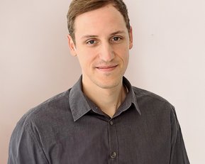 Ein junger Mann mit kurzen, braunen Haaren trägt ein graues Hemd und steht vor einer hellen Wand. Er lächelt leicht und schaut direkt in die Kamera.