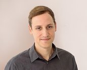 Ein junger Mann mit kurzen, braunen Haaren trägt ein graues Hemd und steht vor einer hellen Wand. Er lächelt leicht und schaut direkt in die Kamera.