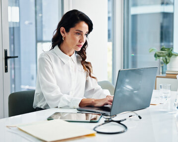 Eine Frau in einem weißen Hemd arbeitet konzentriert an einem Laptop in einem modernen Büro. Auf dem Tisch liegen ein Tablet und Schreibmaterial.