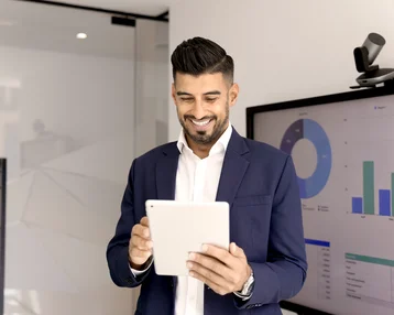 Ein Mann in einem Anzug hält ein Tablet und lächelt, während er vor einem Bildschirm mit Diagrammen und Grafiken steht.