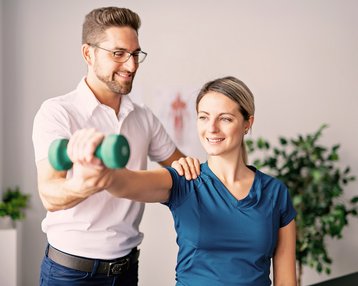 In einer Ergotherapie Sitzung hilft ein Mann einer Frau beim Heben einer Hantel während einer Übungssitzung. Die Frau lächelt, während der Mann sie an der Schulter unterstützt.