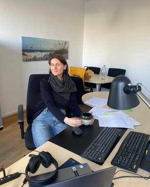 Frau mit einem schwarzen Schal sitzt an einem Schreibtisch und hält eine Tasse in der Hand, umgeben von Unterlagen und Büromaterial.