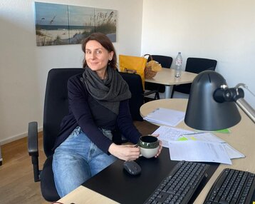 Frau mit einem schwarzen Schal sitzt an einem Schreibtisch und hält eine Tasse in der Hand, umgeben von Unterlagen und Büromaterial.
