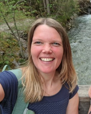 Eine Frau mit langen, blonden Haaren lächelt in die Kamera, während sie an einem Flussufer steht, umgeben von Bäumen.