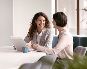 Zwei Frauen sitzen an einem Tisch, eine von ihnen lächelt und hält ein Dokument, während die andere aufmerksam zuhört. Ein Laptop und eine Tasse stehen auf dem Tisch.