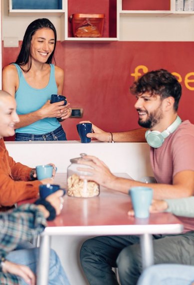 Eine Gruppe junger Menschen sitzt entspannt an einem Tisch und unterhält sich bei Kaffee oder Tee. Eine weitere Person steht lächelnd hinter einer Theke mit der Aufschrift „free food“. Die Atmosphäre wirkt freundlich und einladend, wie in einem Gemeinschaftsraum oder sozialen Treffpunkt.