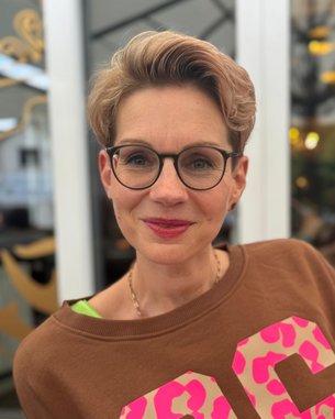 Frau mit kurzen, blonden Haaren und Brille trägt einen braunen Pullover mit großen, pinken Zahlen. Sie lächelt und steht vor einem hellen Hintergrund mit unscharfen Details.
