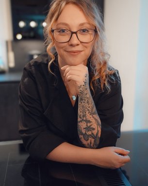 Eine Person mit Brille und tätowiertem Arm lächelt, während sie auf einem Tisch in einer modernen Küche sitzt.