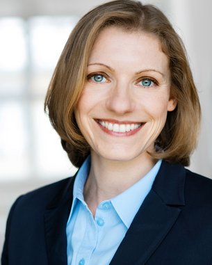 Eine lächelnde Frau mit blonden Haaren trägt einen dunklen Blazer und ein hellblaues Hemd. Der Hintergrund ist unscharf und hell, was den Fokus auf ihr Gesicht lenkt.