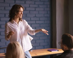 Eine Frau in einem weißen Hemd spricht und gestikuliert vor einem Publikum, das ihr aufmerksam zuhört. Im Hintergrund sind eine blaue Wand und ein Tisch mit Notizmaterialien sichtbar.