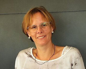 Frau mit kurzen, blonden Haaren und Brille, trägt ein helles Oberteil mit Muster und lächelt freundlich in die Kamera.