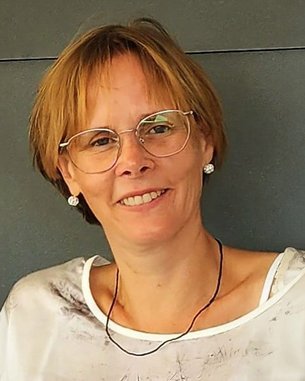 Frau mit kurzen, blonden Haaren und Brille, trägt ein helles Oberteil mit Muster und lächelt freundlich in die Kamera.