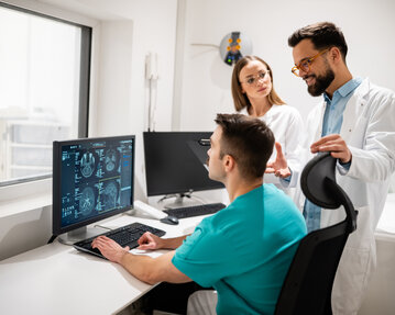 Drei medizinische Fachkräfte besprechen CT-Scans an einem Computerbildschirm in einem modernen Behandlungsraum.