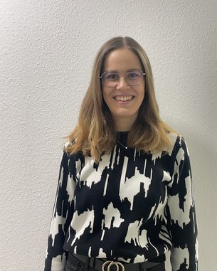 Eine Person mit langen, braunen Haaren trägt einen schwarz-weißen Pullover mit grafischem Muster und eine schwarze Hose mit einem Gürtel. Sie steht vor einer hellen Wand und lächelt.