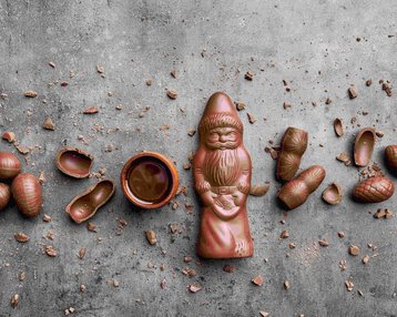 Ein Schokoladenfigur eines Weihnachtsmanns umgeben von Schokoladeneiern und einer kleinen Schüssel mit Schokolade auf einem grauen Untergrund.