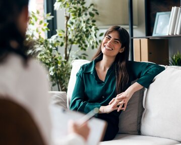 Eine Frau mit langen Haaren sitzt auf einem Sofa, lächelt und spricht mit einer anderen Person, die nicht sichtbar ist.
