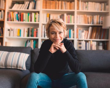 Eine Person mit blonden, kurzen Haaren sitzt auf einem Sofa, lächelt und stützt ihr Kinn auf die Hände. Im Hintergrund sind Bücherregale mit zahlreichen Büchern sichtbar.