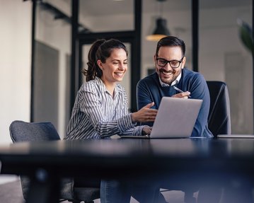Zwei Personen sitzen an einem Tisch und schauen auf einen Laptop. Die Frau zeigt auf den Bildschirm, während der Mann lächelt und aufmerksam zuhört. Beide scheinen an einer Diskussion oder Präsentation beteiligt zu sein.