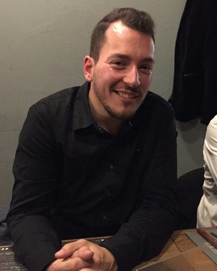 Ein Mann mit kurzem Haar und Bart sitzt an einem Tisch, lächelt und trägt ein schwarzes Hemd. Neben ihm ist eine Person in einem weißen Hemd.