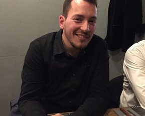 Ein Mann mit kurzem Haar und Bart sitzt an einem Tisch, lächelt und trägt ein schwarzes Hemd. Neben ihm ist eine Person in einem weißen Hemd.