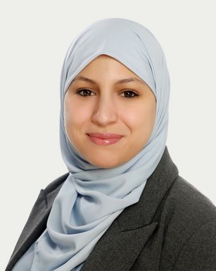 Eine Frau mit einem hellblauen Hijab und einem grauen Blazer lächelt in die Kamera. Der Hintergrund ist neutral und hell.