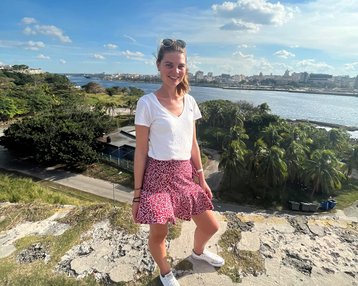 Eine junge Frau steht auf einer Mauer mit Blick auf eine Stadtlandschaft und einen Fluss. Sie trägt ein weißes T-Shirt, einen gemusterten Rock und Sneakers. Im Hintergrund sind Bäume und Gebäude sichtbar.