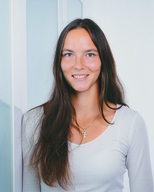 Eine Frau mit langen, braunen Haaren steht lächelnd neben einer Glaswand. Sie trägt ein helles Oberteil und hat eine Kette um den Hals.