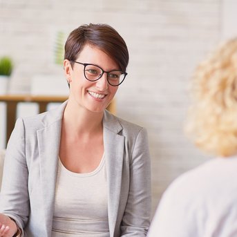Eine lächelnde Frau mit Brille in einem grauen Blazer sitzt auf einem Sofa und spricht mit einer anderen Person.
