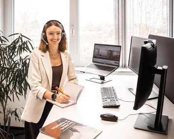 Eine Frau mit Headset sitzt an einem Schreibtisch und arbeitet am Computer, während sie in einem modernen Büro umgeben von Pflanzen ist.