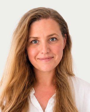 Eine Frau mit langen, lockigen Haaren und einem freundlichen Gesichtsausdruck trägt ein weißes Hemd. Der Hintergrund ist neutral.