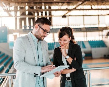 BWL und Sportmanagement Bachelor Ein Mann und eine Frau stehen in einem Sportstadion und betrachten gemeinsam ein Tablet. Beide scheinen in ein Gespräch vertieft zu sein, während im Hintergrund leere Sitzplätze sichtbar sind.