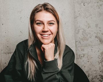 Eine lächelnde Frau mit langen, blonden Haaren sitzt in einem schwarzen Stuhl und stützt ihr Kinn auf die Hand. Der Hintergrund besteht aus einer grauen Wand.