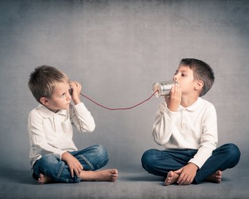 Zwei Jungen sitzen auf dem Boden und kommunizieren über eine selbstgemachte Dose mit Schnur. Einer hält die Dose ans Ohr, während der andere in die Dose spricht. Beide tragen weiße Hemden und Jeans.